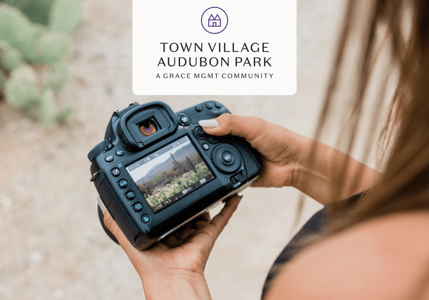 Person holding a DSLR camera outdoors, capturing a landscape with a logo card for Town Village Audubon Park above the frame.